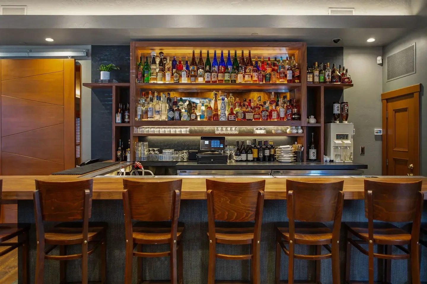 Interior bar at Matsuhisa Aspen, showcasing bottles and wooden chairs.